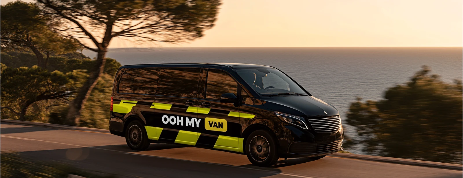 Van noir en Grand côté jaune-vert marqué OOH MY VAN, sur une route du littoral au coucher du soleil
